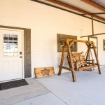 A covered patio with a white door, two black-trimmed windows, a wooden porch swing, a welcome mat, and a wooden sign leaning against a white brick wall. The floor is concrete and part of a metal roof is visible.