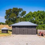 A small brown shed and a larger brown storage building with metal roofs sit on a gravel lot, surrounded by trees, grass, and a wooden fence under a clear blue sky.