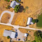 Aerial view of a rural property with several buildings, a pond, trees, parked vehicles, driveways, and a surrounding grassy landscape adjacent to a road.