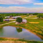 A small farmhouse with a gray roof sits by a pond, surrounded by green fields, trees, and farm buildings under a colorful sky, with a scenic rural landscape in the background.
