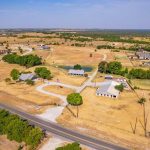 Aerial view of a rural residential area with large homes, barns, scattered trees, and open dry fields along a winding road under a clear blue sky.
