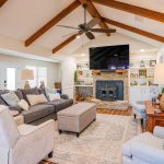 A spacious living room with vaulted ceiling and exposed wooden beams, featuring a gray sectional sofa, armchairs, a stone fireplace with a large TV above, built-in shelves, and soft, neutral decor.