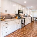 Modern kitchen with white cabinets, stainless steel appliances, a marble countertop, and a dark tiled backsplash. The space features wood flooring and a dining area with a large "M" on the back wall.
