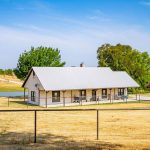 A single-story house with a metal roof sits on dry, yellow grass, surrounded by a black wire fence. Trees and a small pond are visible in the background under a clear blue sky.