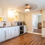 A spacious kitchen with white cabinets, wood flooring, a stove, oven, microwave, and coffee maker. The walls have beige tile backsplash, and a ceiling fan is overhead. An open doorway leads to another room.