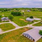 Aerial view of a rural property with multiple houses, winding driveways, green lawns, scattered trees, a pond, and several parked vehicles, surrounded by open fields and rolling hills.