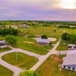 Aerial view of a rural property with several buildings, green lawns, winding roads, scattered trees, and distant houses under a partially cloudy sky at sunset.