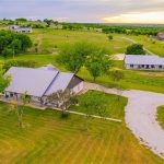 Aerial view of a ranch with a main house, a barn, trees, and a curved gravel driveway, surrounded by green fields and neighboring properties under a partly cloudy sunset sky.