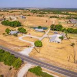 Aerial view of a rural property with several buildings, trees, and fenced areas, surrounded by open fields and scattered houses, adjacent to a two-lane road under a clear sky.