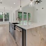 Modern kitchen with a large marble island, stainless steel appliances, light wood floors, pendant lights, and large windows overlooking a fenced backyard. The kitchen opens into a spacious living area with a fireplace.