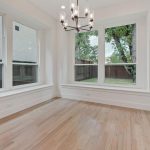 Bright, empty room with light wood flooring, large windows providing a view of trees and grass outside, a glass door to the right, and a modern chandelier hanging from the ceiling.