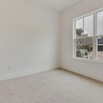 Empty room with light-colored carpet, white walls, and a large window showing a view of neighboring houses and a street outside. The room has a closed white door and appears clean and bright.