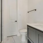 Modern bathroom with a glass shower enclosure, white toilet, gray vanity with white countertop, dark faucet, and towel bar. The floor and shower have white marble-like tiles. Walls are light-colored.
