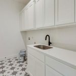 A modern laundry room with white cabinets, a stainless steel sink, a black faucet, and patterned tile flooring. Two paint cans are on the floor near the wall. The space is clean and bright.