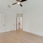 A bright, empty room with light wood flooring, white walls, a ceiling fan with light, and multiple doors leading to other areas, including a bathroom.