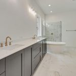 Modern bathroom with double sinks on a long vanity, gold fixtures, a freestanding white bathtub, glass-enclosed shower, marble floors, and white walls in a bright, spacious setting.