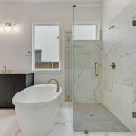 Modern bathroom with a freestanding white tub, glass-enclosed shower, double sink vanity with dark cabinets, gold fixtures, and two windows; white marble floors and walls enhance the bright, clean look.