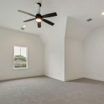 A spacious, empty room with white walls, a gray carpet, a ceiling fan with light, recessed lighting, and a single window letting in natural light. The room has high, sloped ceilings.