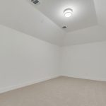 A clean, empty room with white walls, beige carpet, a ceiling light fixture, and angled ceilings. There are air vents on the ceiling and electrical outlets along the lower walls.