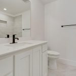 A modern, minimalistic bathroom with white cabinets, a large mirror, black faucet and handles, a toilet, and tiled walls and floors in neutral tones. The shower area is visible in the mirror.