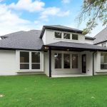 A modern two-story house with white brick exterior and black trim, featuring large windows and a covered back patio, surrounded by a wooden fence and green lawn under a partly cloudy sky.