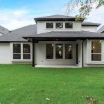 Back view of a modern two-story house with white brick exterior, covered patio, large windows, and a fenced backyard with green grass. Neighboring houses are visible on both sides.