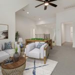 A modern living room with gray sofas, patterned pillows, a round wooden coffee table, and wall art of coastal scenes; a pool table sits in the back near a staircase and hallway.