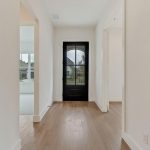 A bright, modern hallway with light wood floors and white walls features a large black-framed glass front door, allowing natural light to enter. Rooms are visible on either side, with windows overlooking a suburban yard.