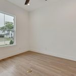An empty room with light-colored walls, a large window letting in natural light, wooden flooring, and a small round floor outlet near the center.