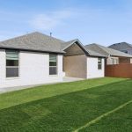 Single-story white brick house with a covered patio, fenced backyard, and green grass, situated in a suburban neighborhood under a clear blue sky.