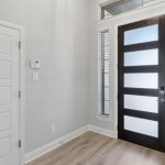 Modern entryway with light wood flooring, a white interior door on the left, a dark front door with frosted glass panels, and tall windows with blinds on either side. Walls are light gray and the space is well-lit.