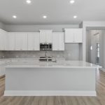 Modern kitchen with white cabinets, marble backsplash, stainless steel microwave, and a large white island with sink. Light wood flooring and recessed ceiling lights create a bright, spacious atmosphere.