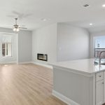 Open-concept living area with light wood flooring, white walls, a modern kitchen island with a sink, built-in fireplace, large windows, and glass doors leading to a fenced backyard.