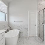 Modern bathroom with white cabinets, a freestanding bathtub, marble flooring and walls, a glass-enclosed shower, and a large window with blinds letting in natural light.