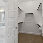A spacious walk-in closet with built-in shelves and hanging rods, viewed from the doorway. The closet has carpeted flooring and connects to a white bathroom with a bathtub and vanity visible.
