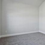 Empty room with light gray walls, large windows on the left, gray carpet, and an attached bathroom with tan tile flooring and a white vanity. Natural sunlight brightens the space.