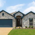 Single-story suburban house with a two-car garage, gray brick exterior, dark trim, manicured lawn, flower beds, under a partly cloudy sky.