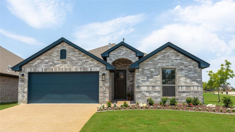 Single-story suburban house with a two-car garage, gray brick exterior, dark trim, manicured lawn, flower beds, under a partly cloudy sky.