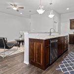 Modern open-concept kitchen and living area with dark wood cabinets, granite countertops, stainless steel appliances, a fireplace, armchair, coffee table, and ceiling fan. Brown tile flooring throughout.