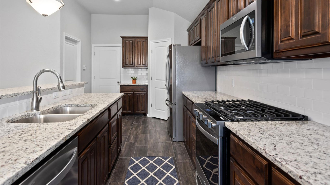Modern kitchen with dark wood cabinets, stainless steel appliances, granite countertops, a double sink, gas stove, and white tile backsplash. A small geometric rug lies on the dark wood floor.