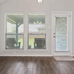 Bright room with three large windows and a glass door leading outside, featuring wood-look tile flooring. Adjacent room with a bed is visible through an open doorway on the right.