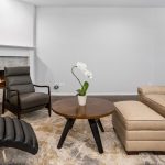 A modern living room with a beige sofa, matching ottoman, black armchair, round wooden coffee table with a white orchid, black chaise lounge, fireplace, and neutral walls.