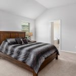 A modern bedroom with a wooden bed frame, black bedding, two windows with blinds, a wooden dresser with a TV, beige carpet, and a door leading to a bright adjoining bathroom.