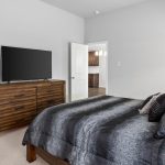 A modern bedroom with a wooden dresser holding a flat-screen TV, a bed with dark patterned bedding and pillows, light carpet, and an open door leading to another room with wooden cabinets.