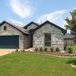 Single-story brick house with a dark garage door, well-maintained front yard, landscaped garden, young tree, and bright blue sky with scattered clouds. Neighboring homes and a suburban street are visible.