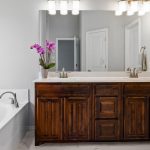 Modern bathroom with a wooden double-sink vanity, large mirror, wall-mounted lights, white bathtub, towel ring, and a purple orchid plant on the counter. White walls and doors complete the clean, bright space.
