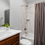 A modern bathroom with a white sink, wooden vanity, potted plant, toilet, and a bathtub with marble-tiled walls and a gray shower curtain. Towels hang on a rack beside the sink.