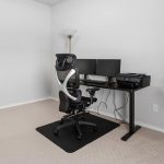 A minimalist home office with a black desk, dual monitors, an office chair, a lamp, and a potted plant in the corner. The room has light-colored walls and carpet.