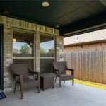 A covered patio with two wicker chairs and a small table sits outside a brick house. A doormat lies by a glass door, and a wooden fence encloses the grassy backyard.