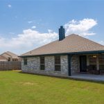 A single-story house with gray brick exterior and a covered back patio, set in a fenced backyard with a lawn under a partly cloudy sky. Outdoor seating is visible on the patio.